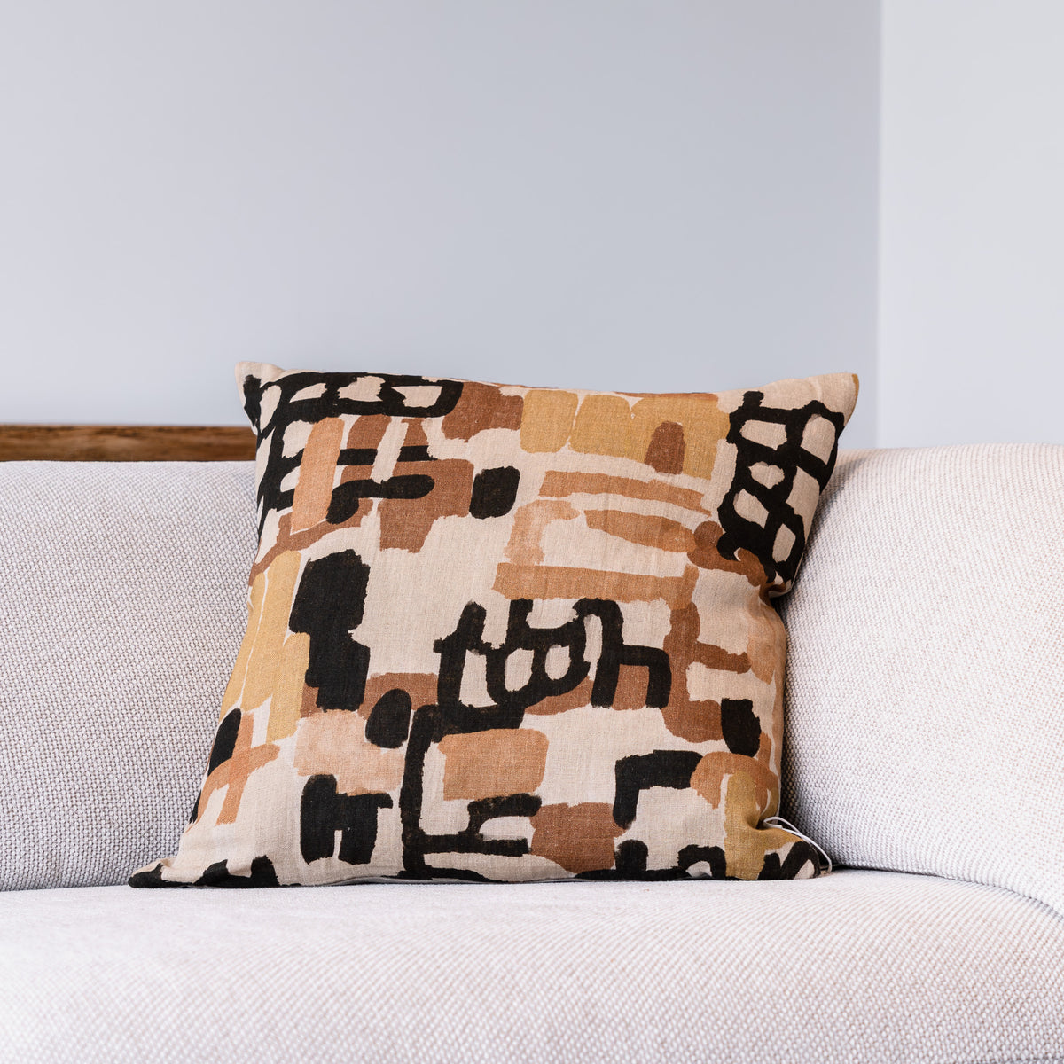 A square throw pillow with an abstract geometric pattern in beige, brown, and black, resting on a light gray textured sofa against a light gray wall.