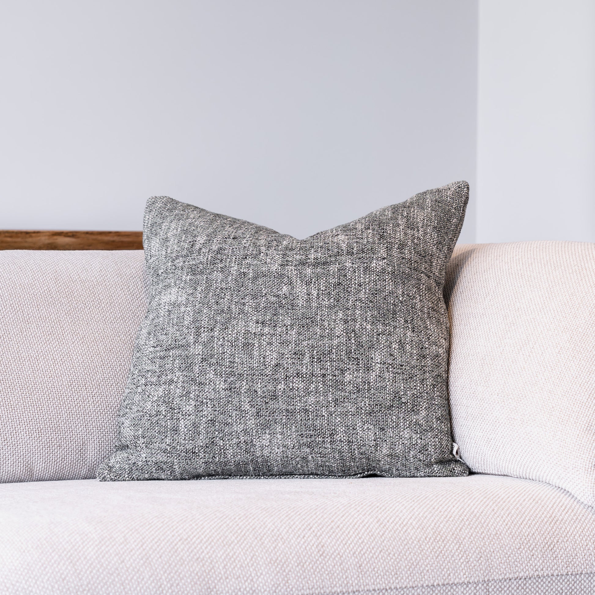 A square gray textured throw pillow resting on a light beige sofa against a neutral gray wall.