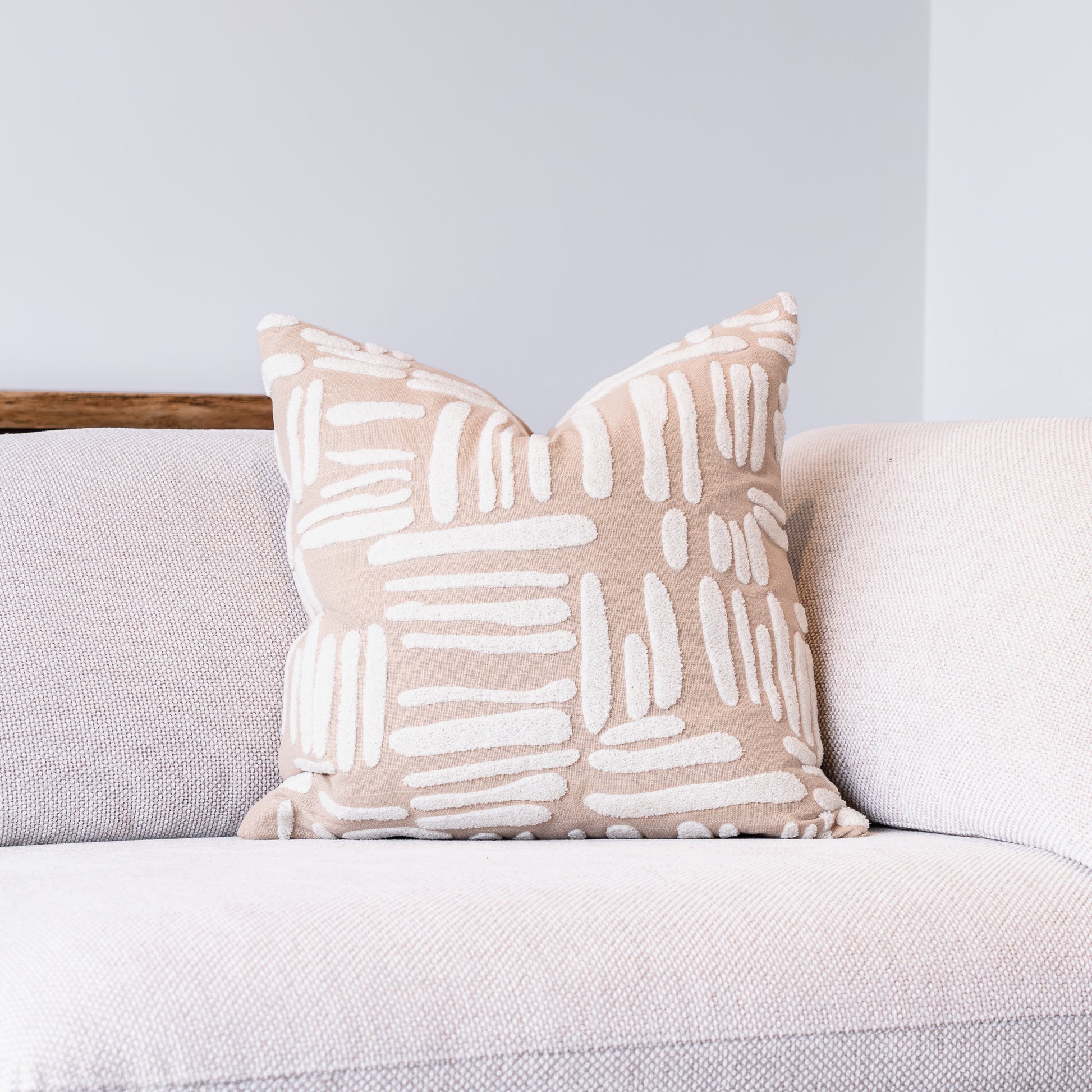 A square throw pillow with a beige and white abstract brushstroke pattern, resting on a light gray textured sofa against a neutral gray wall.