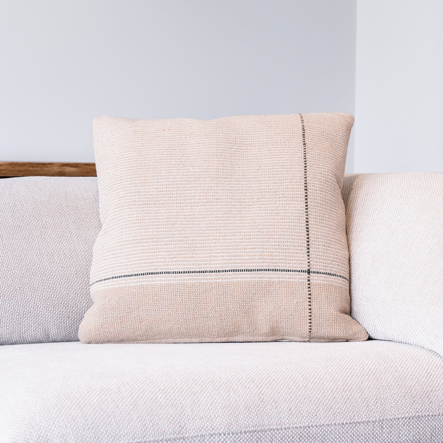 A square beige woven throw pillow with a vertical black stitched line and a horizontal black stripe, resting on a light-colored textured sofa.