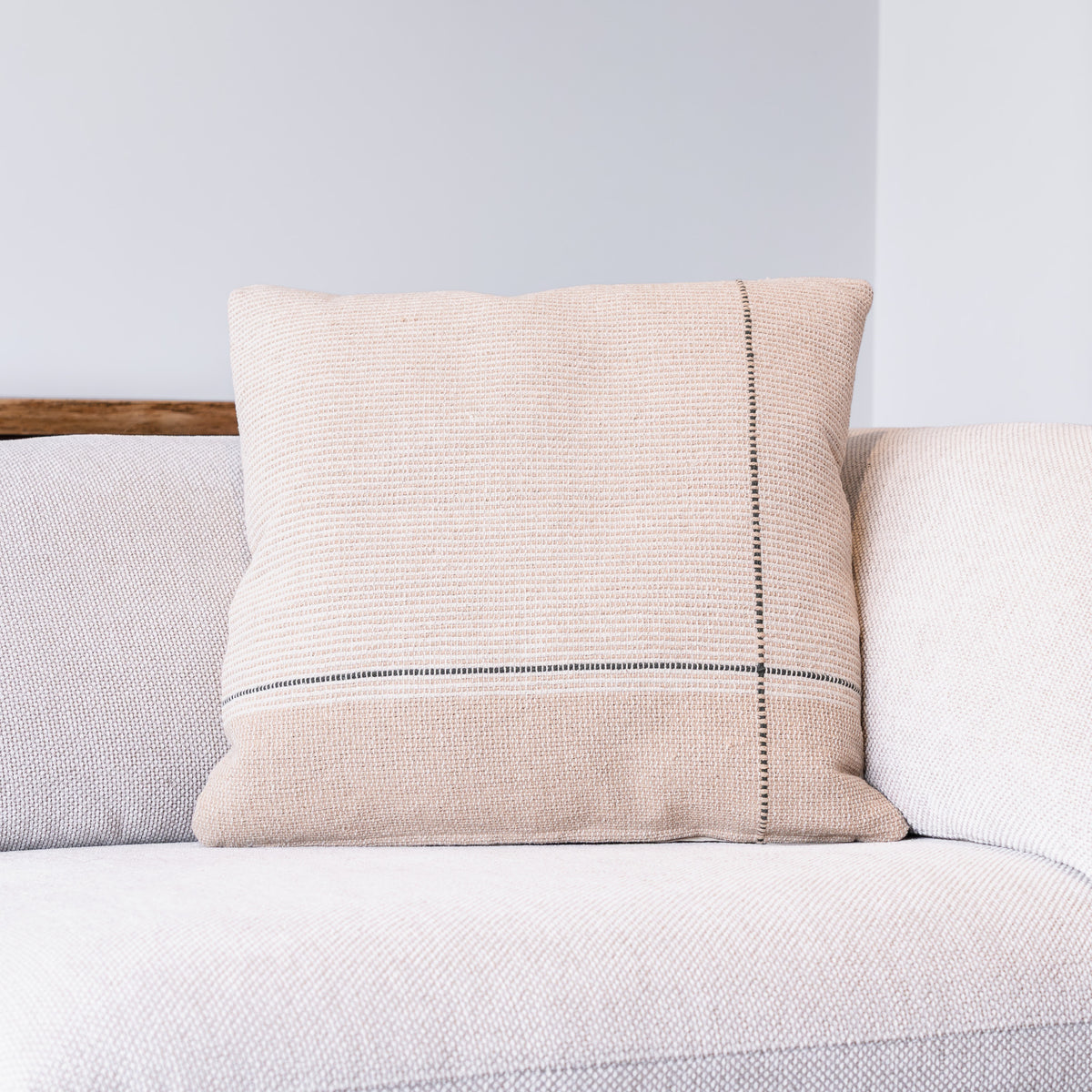 A square beige woven throw pillow with a vertical black stitched line and a horizontal black stripe, resting on a light-colored textured sofa.