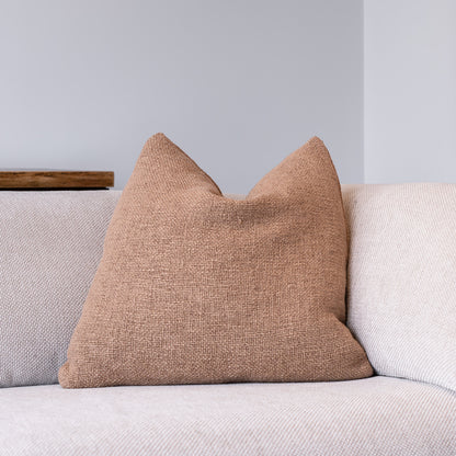 A square throw pillow in a textured tan linen fabric, styled on a light beige sofa against a neutral gray wall.