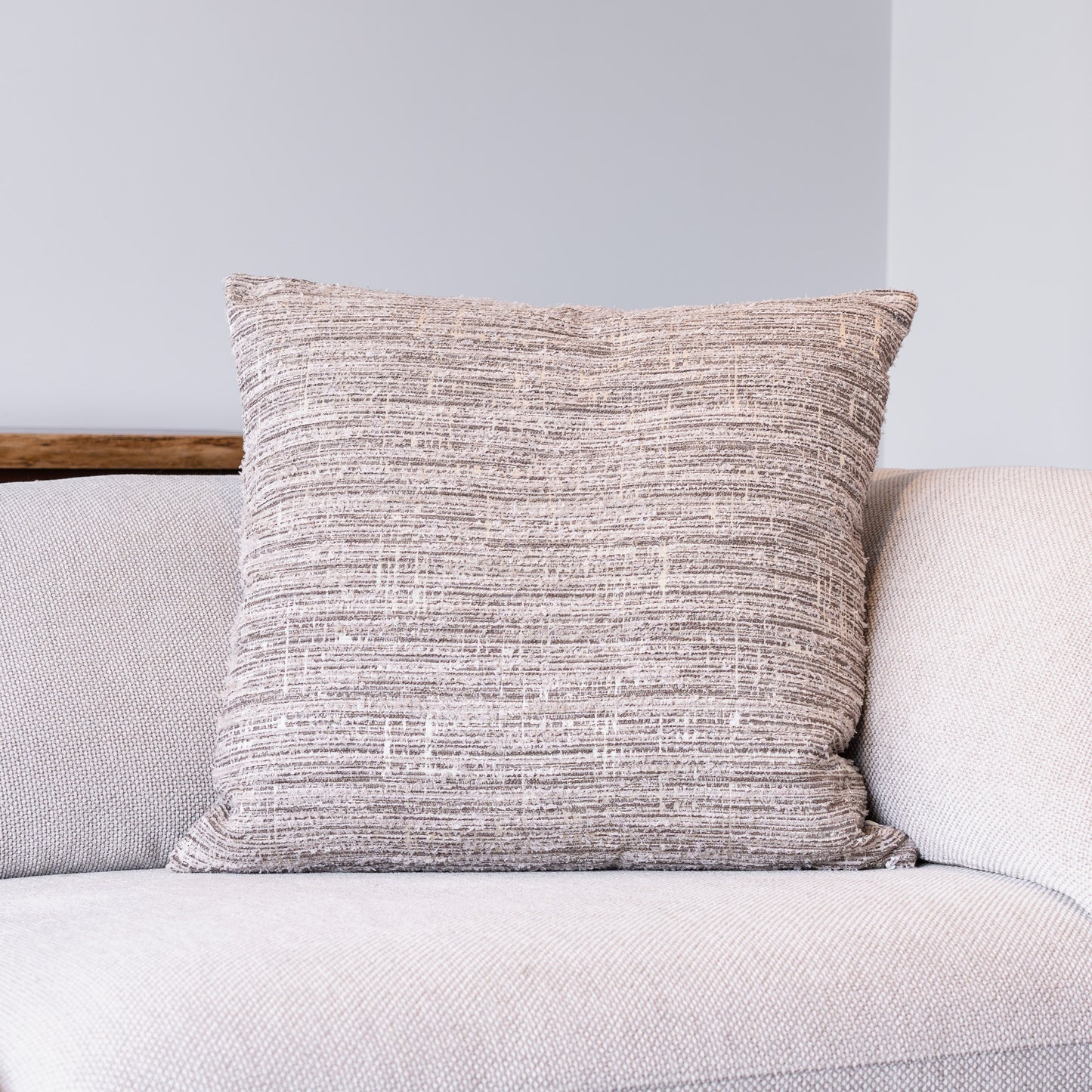 A square throw pillow with a textured, heathered taupe and beige fabric, resting on a light gray sofa against a neutral gray wall.