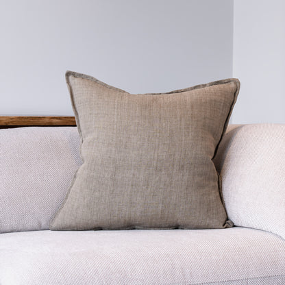 A square beige throw pillow with a textured linen-like fabric and dark gray piped edges, resting on a light gray textured sofa against a neutral gray wall.