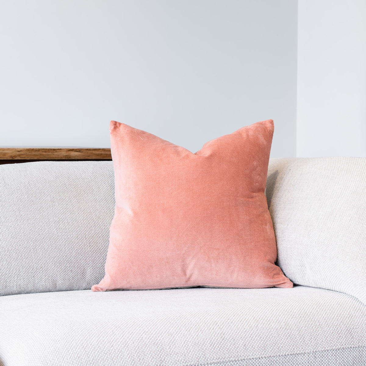 A square coral pink velvet throw pillow resting on a light gray textured fabric sofa against a neutral light gray wall.
