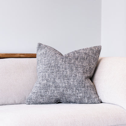 A square throw pillow with a textured gray and white woven pattern, resting on a light beige sofa against a neutral gray wall.