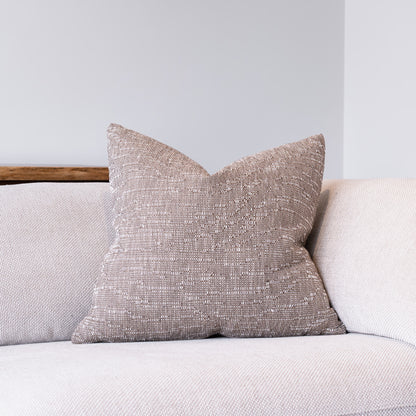 A square throw pillow with a textured, woven taupe fabric sits on a light beige sofa against a neutral gray wall.