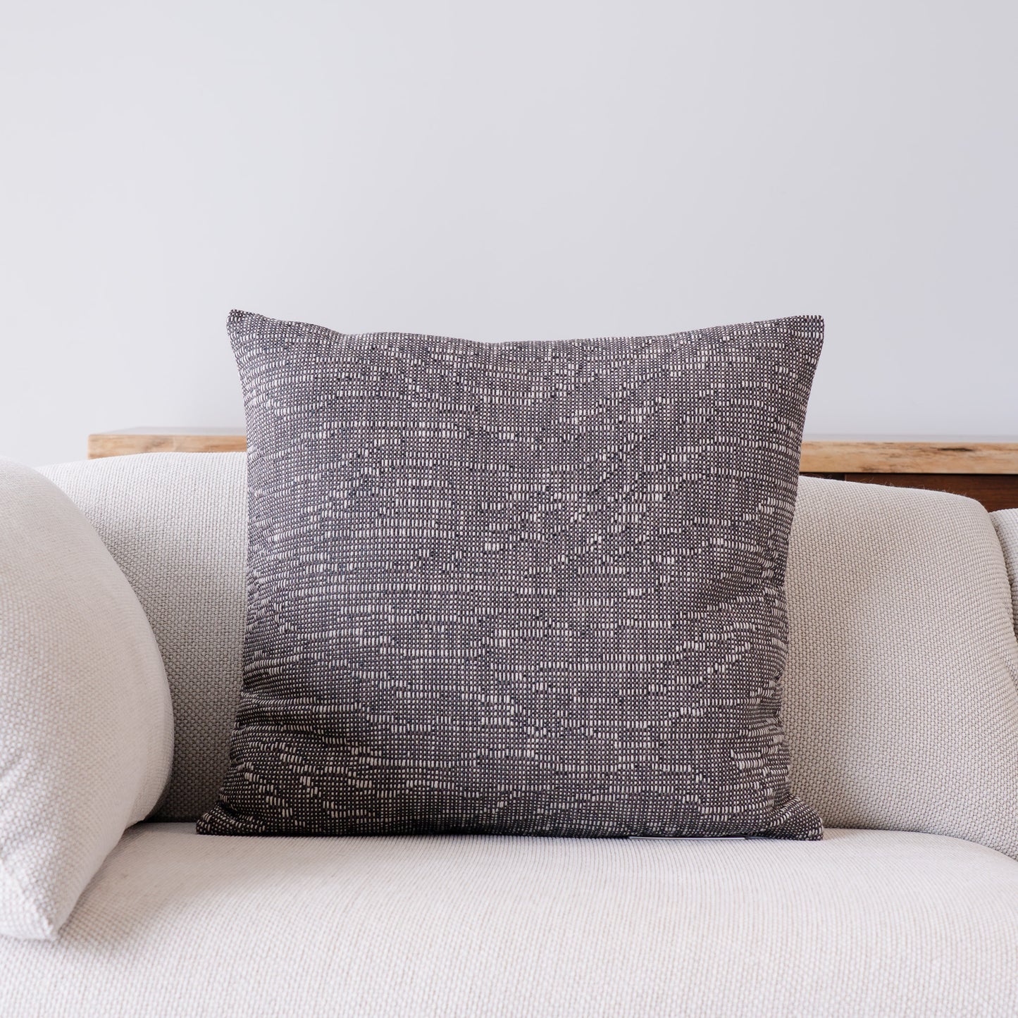 A square throw pillow with a textured, woven charcoal gray and off-white pattern, displayed on a light beige sofa against a neutral wall.
