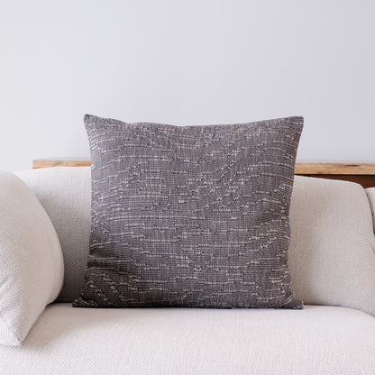 A square throw pillow with a textured, woven charcoal gray and off-white pattern, displayed on a light beige sofa against a neutral wall.