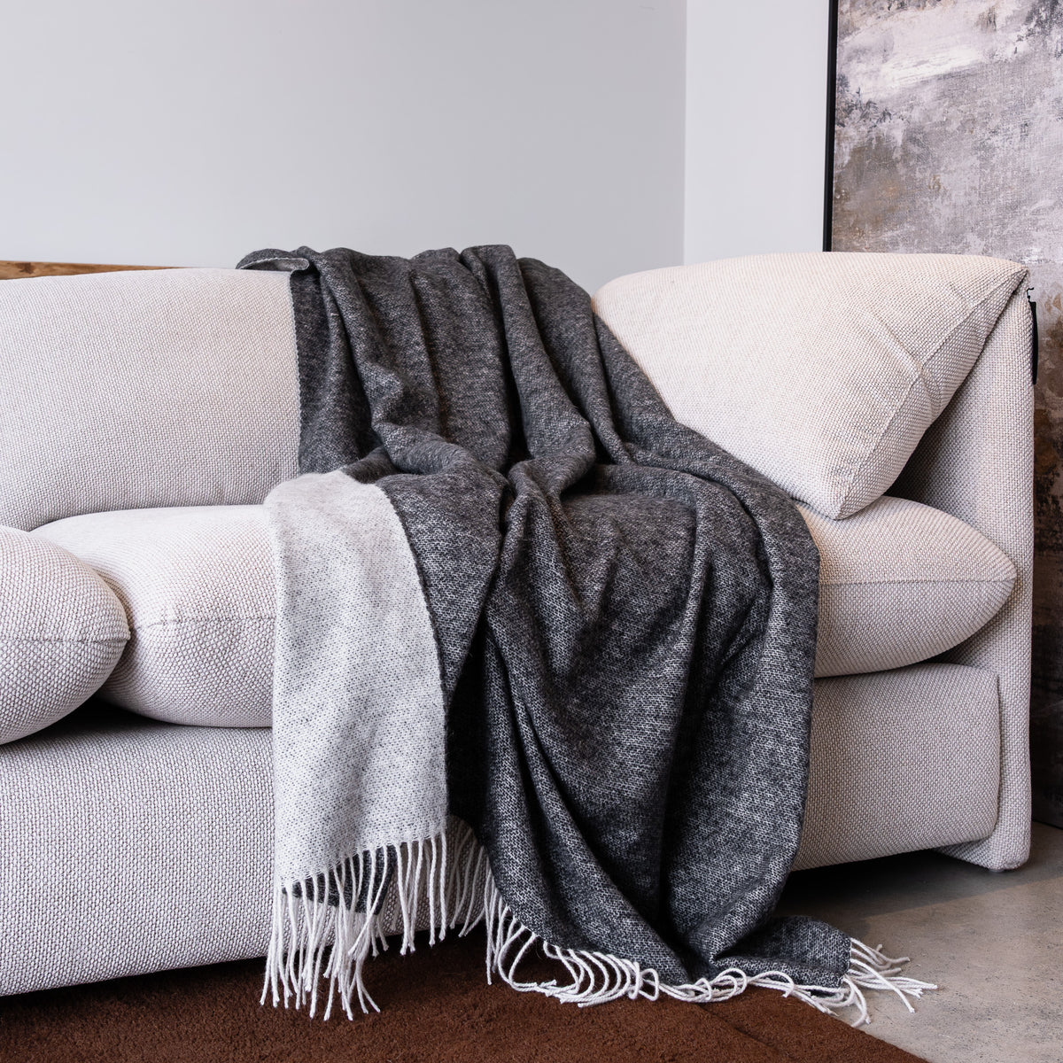 A dark charcoal gray and light beige woven throw blanket with white fringe, draped over a light beige fabric sofa in a modern living room setting.