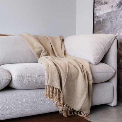 A light beige woven throw blanket with fringed edges draped over a modern light gray sofa in a minimalist room setting.
