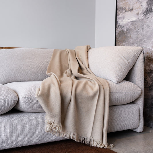 A light beige woven throw blanket with fringed edges draped over a light gray fabric sofa in a modern living room setting.